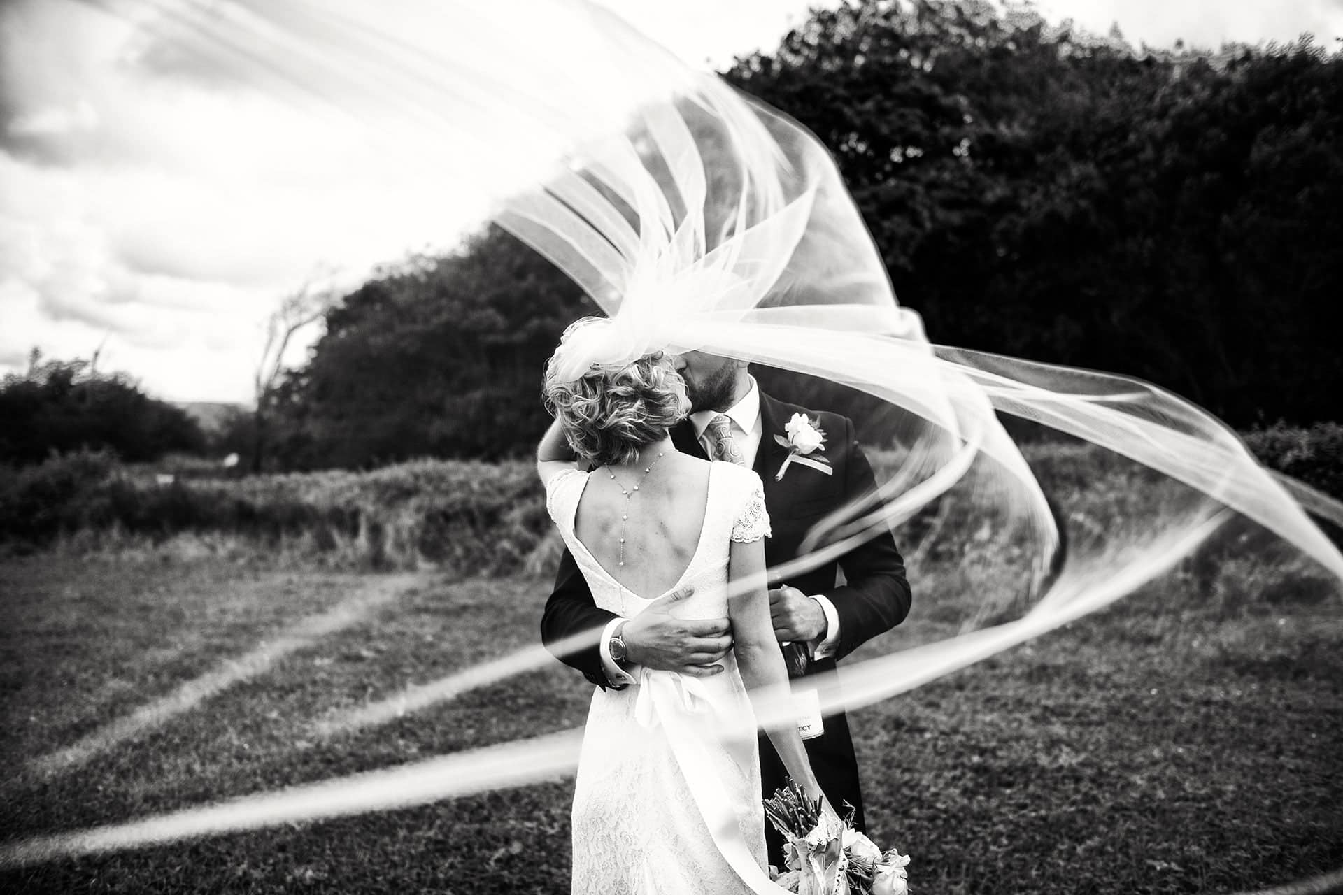 Aberaeron Wedding Photographer in West Wales. Black and white image of the Bride and groom photographed behind a veil swirling in the wind. This photo won an international ISPWP award in the movement and motion section.